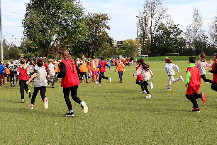 Viele Kinder mit bunten Trikots laufen über einen Sportplatz.