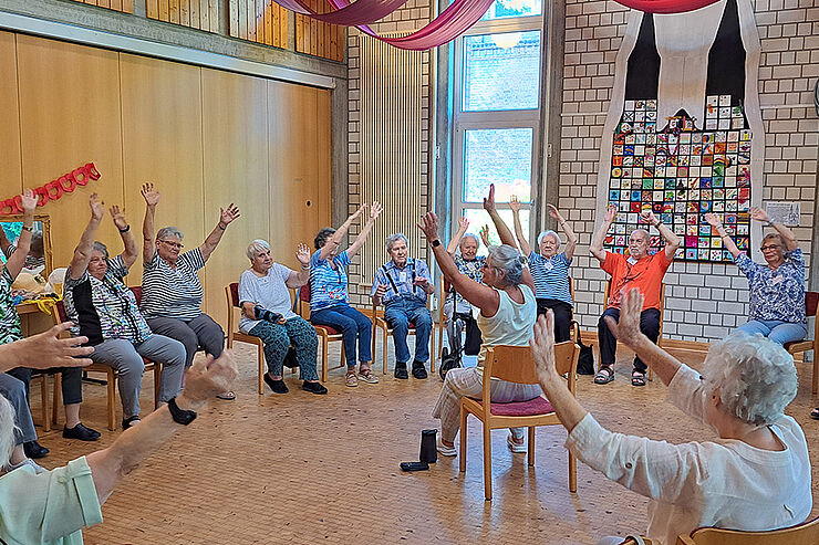 Menschen sitzen im Stuhlkreis und winken mit den Armen.