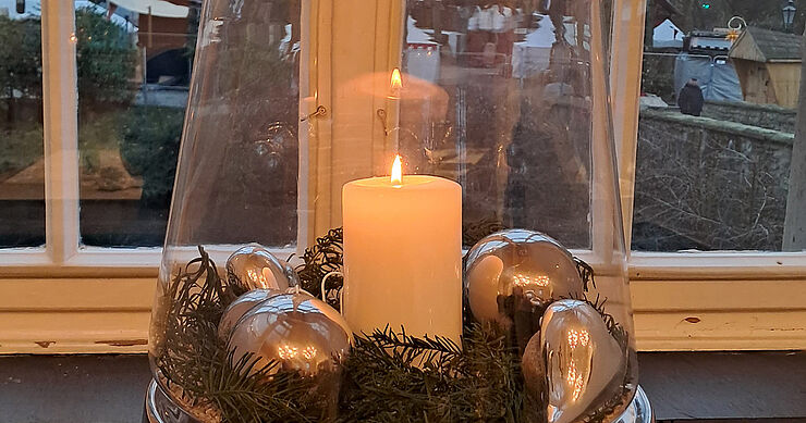 Eine angezündete Kerze im Glas steht im Fenster.