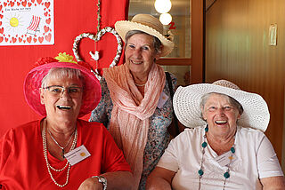 Drei Frauen mit Strohhüten sitzen vor einer roten Wand und lachen.